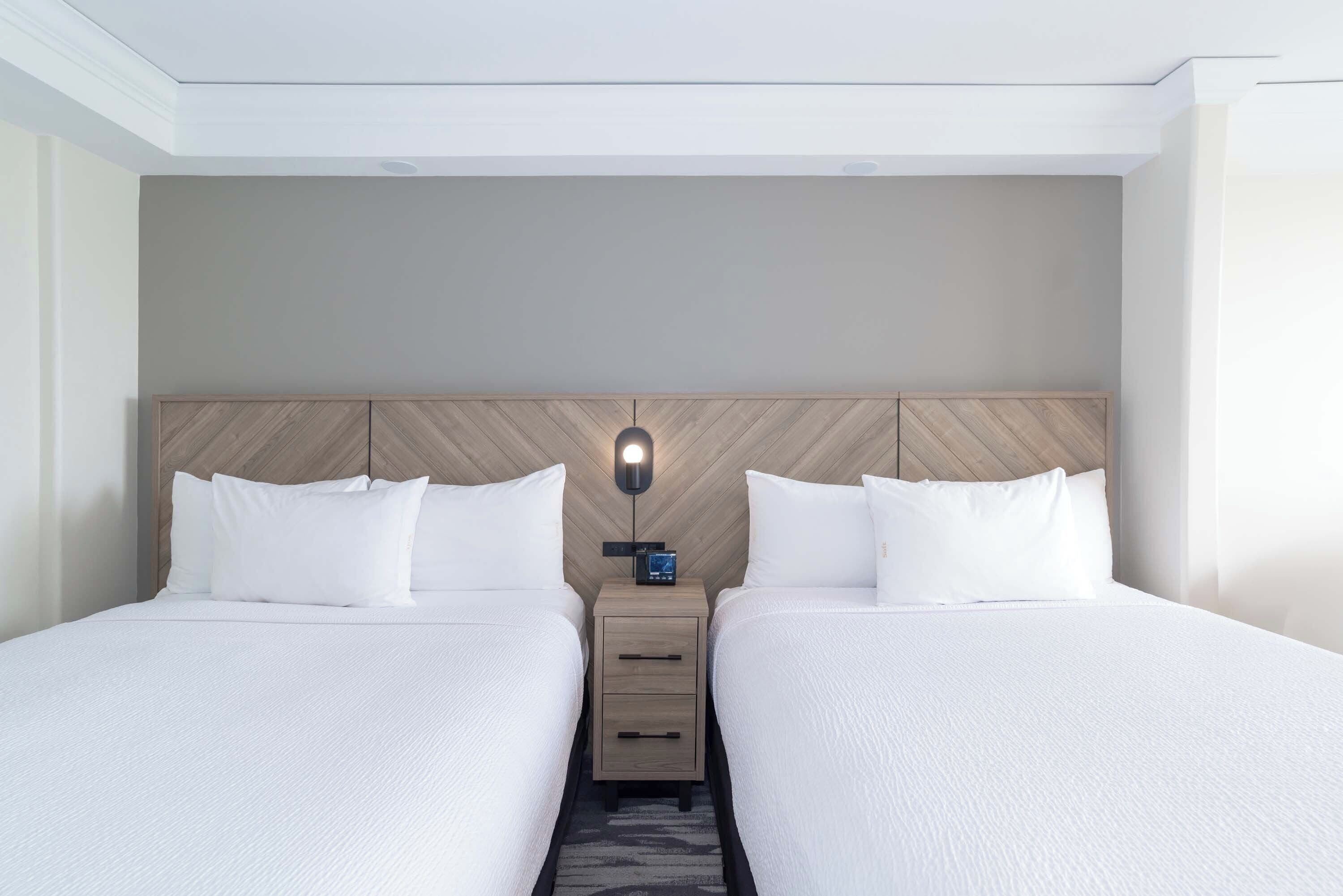 A hotel room with two beds, a wooden headboard, a nightstand with a clock, and a modern light fixture.