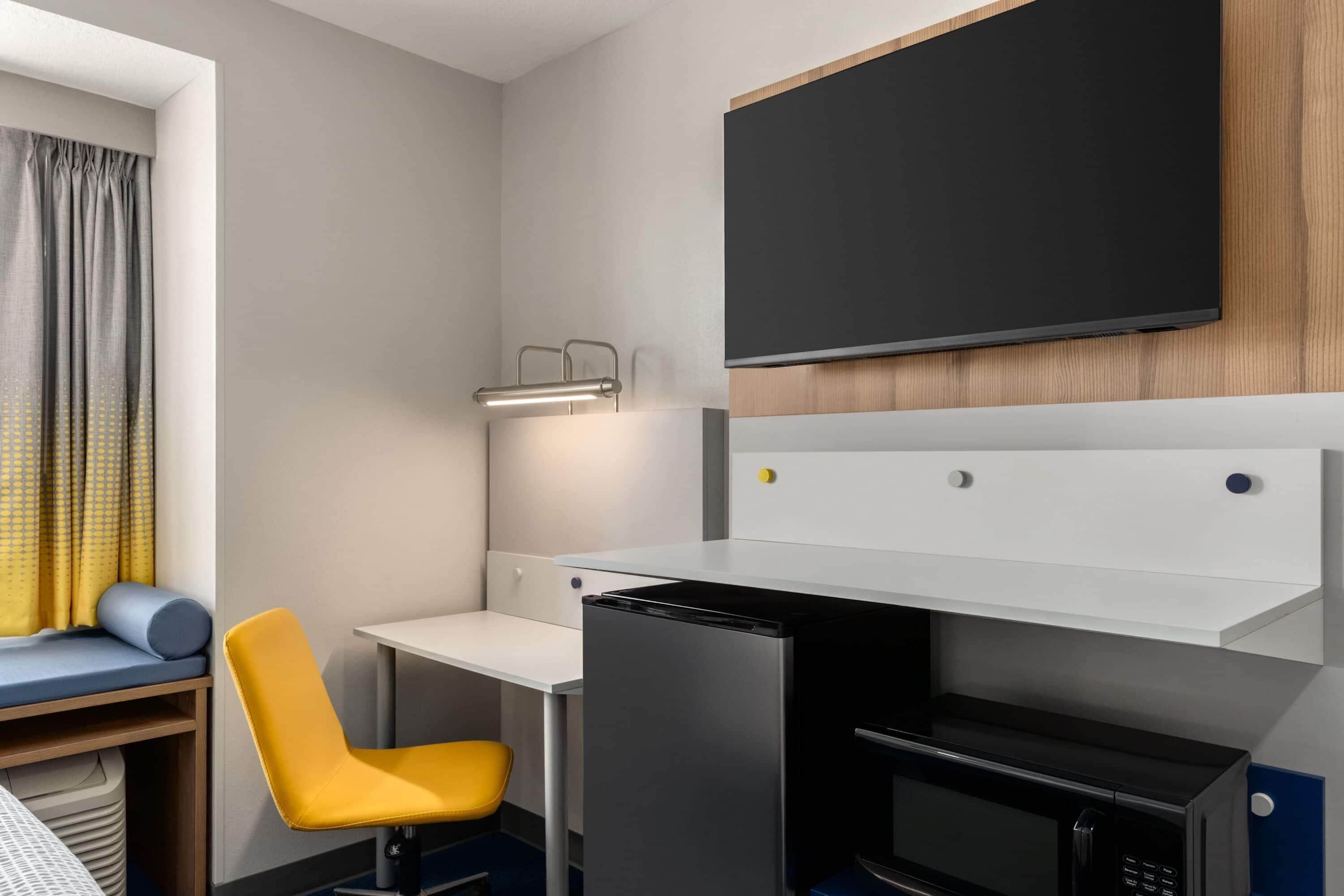 Una habitación de hotel compacta con cama, un escritorio pequeño, microondas, refrigerador, una silla amarilla, televisor y ventana con cortinas.