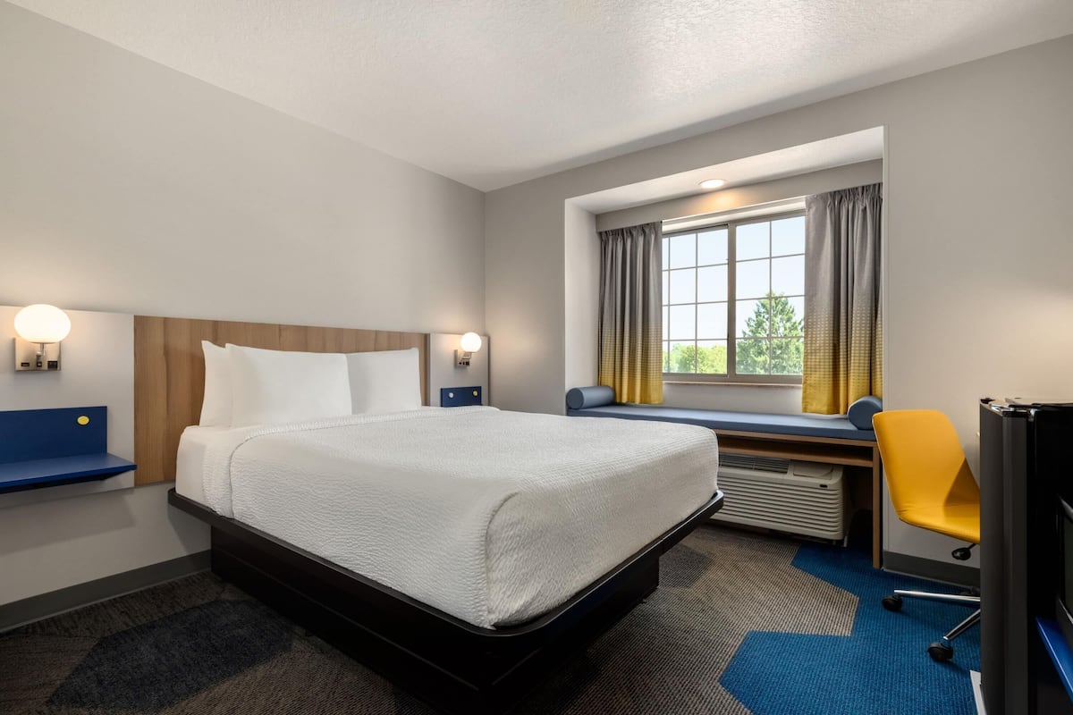 Ein Hotelzimmer mit Bett, Nachttischen, Schreibtisch, Stuhl, einem Fenster mit Vorhängen und Blick ins Grüne.