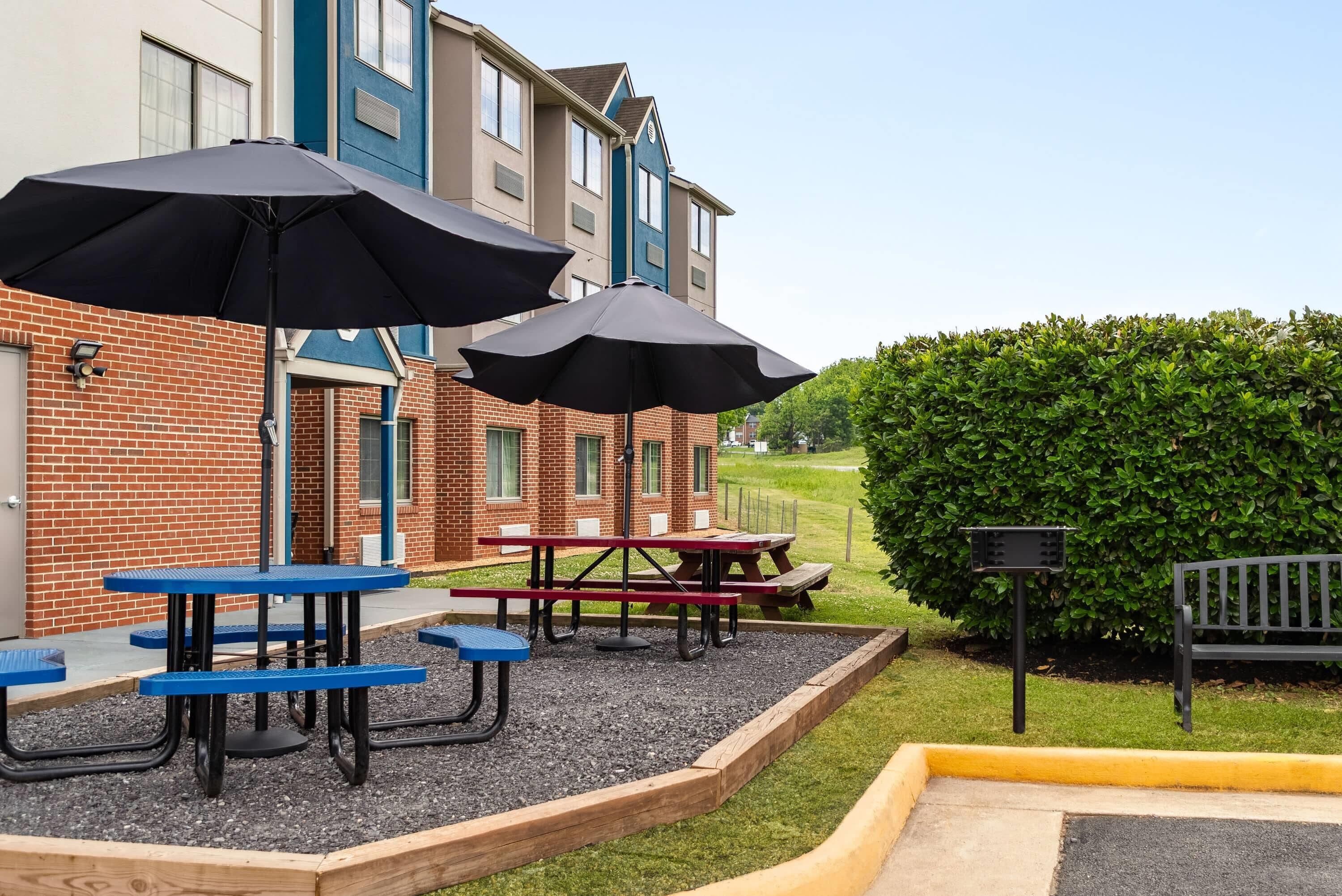 A courtyard with picnic tables and umbrellas, a brick building, and a hedge.
