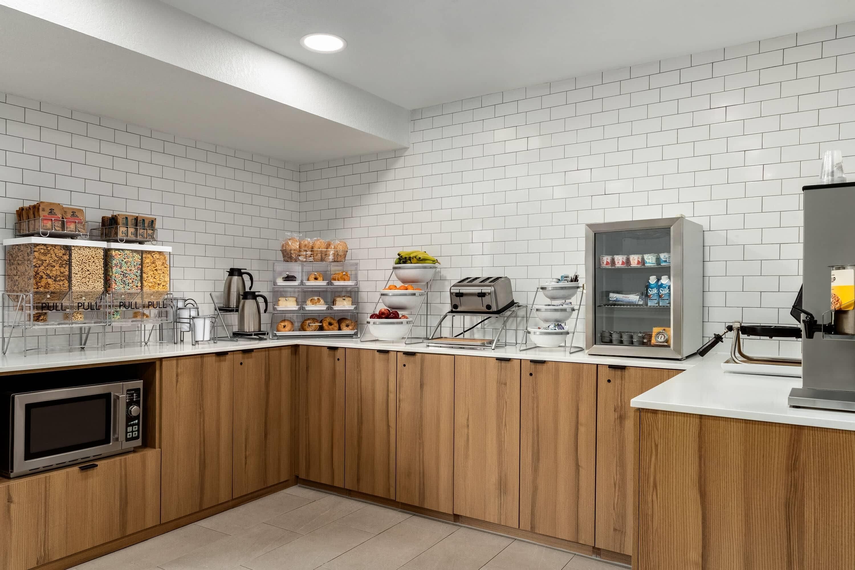Una cocina moderna con paredes de azulejos blancos, armarios de madera, microondas, cafetera, tostadora y refrigerador.