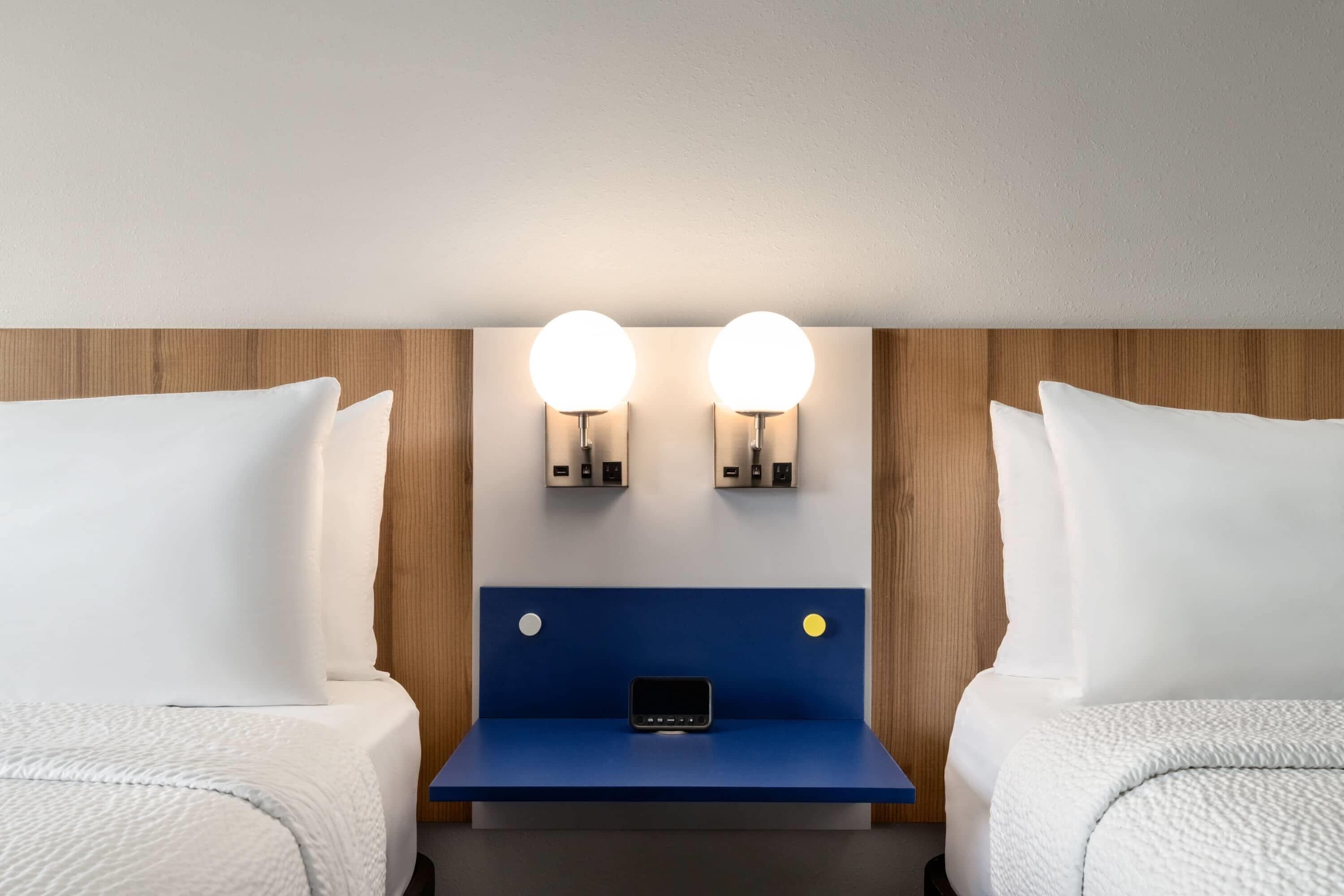 Habitación de hotel con dos camas, una mesita de noche azul y dos lámparas de pared.