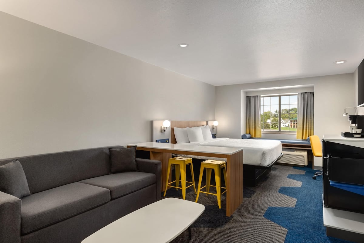 Ein Hotelzimmer mit Bett, Sofa, Schreibtisch mit zwei Hocker, Fenster mit Vorhängen und Blick ins Freie.