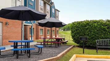 A patio area with picnic tables and umbrellas, a brick building, and a hedge.