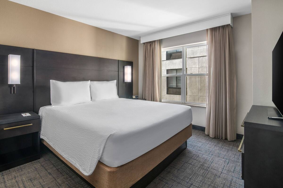 A hotel room with a large bed, a nightstand, a desk, a television, and a window with curtains.