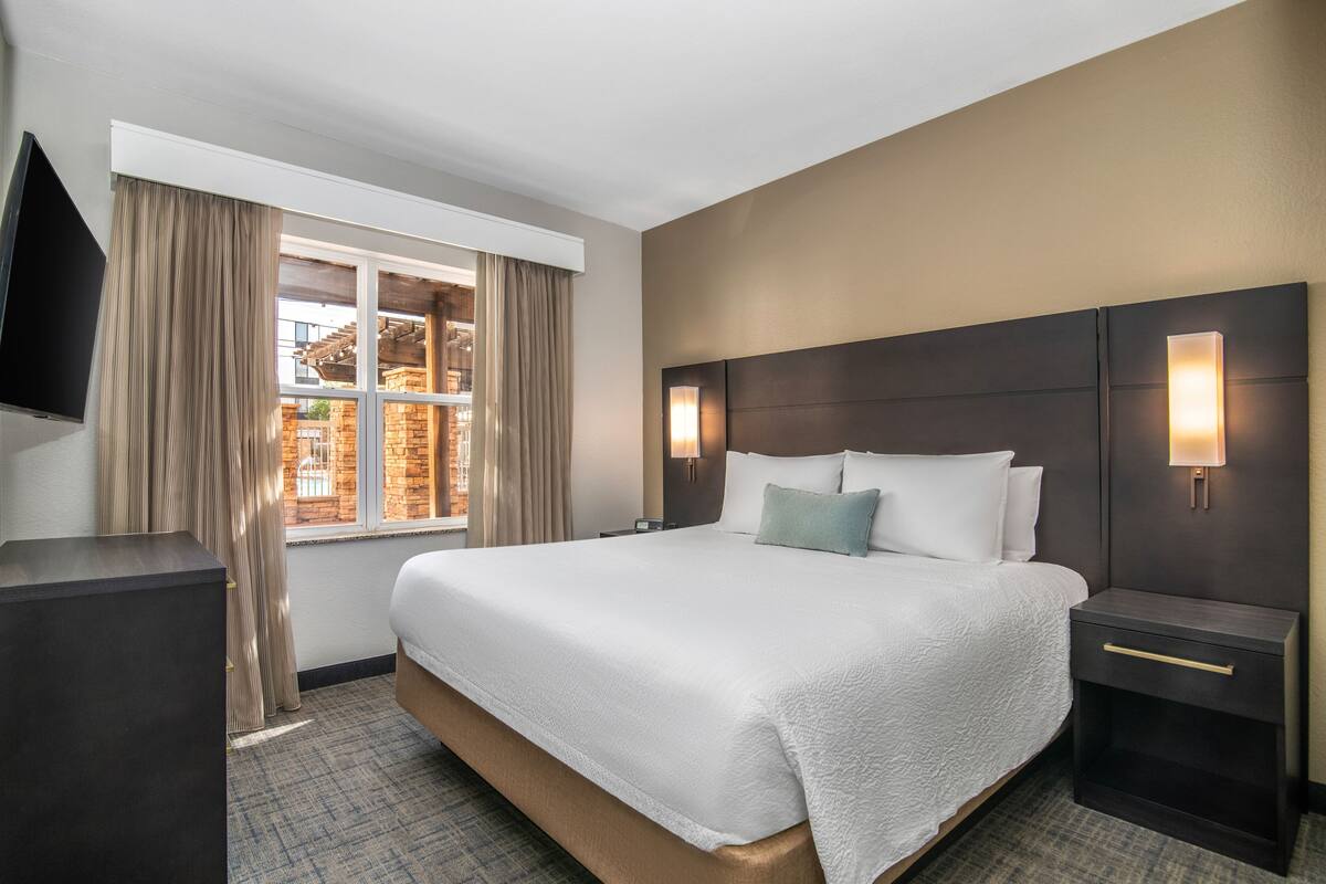 A hotel room with a large bed, a nightstand, a TV, and a window with curtains.