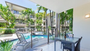 Un balcón con mesa de vidrio, sillas y vista a una piscina y follaje circundante.