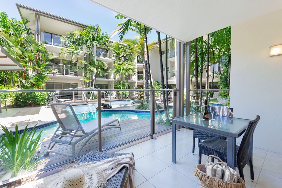 A balcony with a glass table, chairs, and a view of a swimming pool and surrounding greenery.
