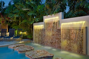 Zona de piscina con muro de piedra y una cascada, rodeada de vegetación exuberante y sillas de descanso.