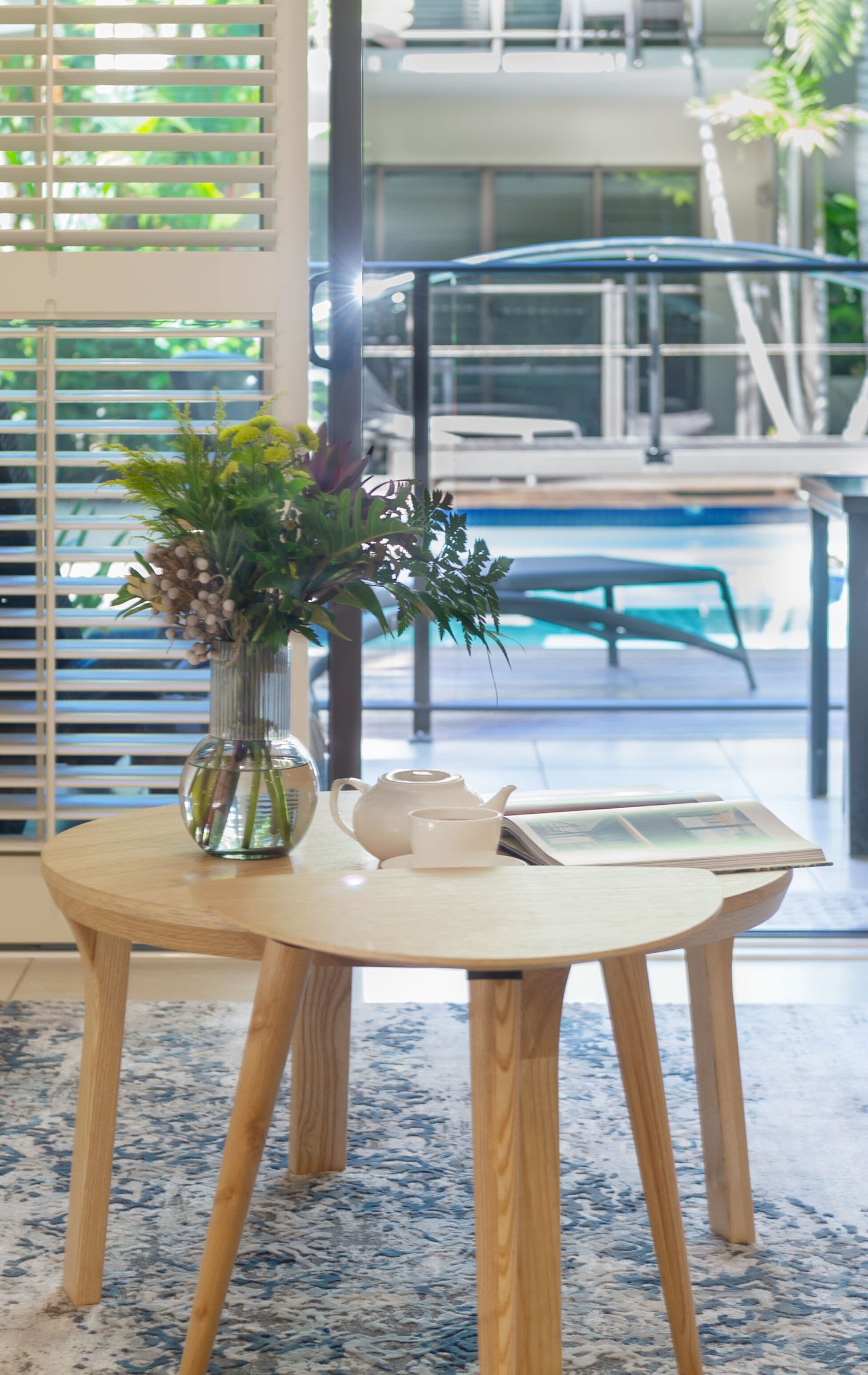 A round wooden table with a clear glass vase containing green plants, a white teapot, and a cup. The table is placed on a blue and white patterned rug.