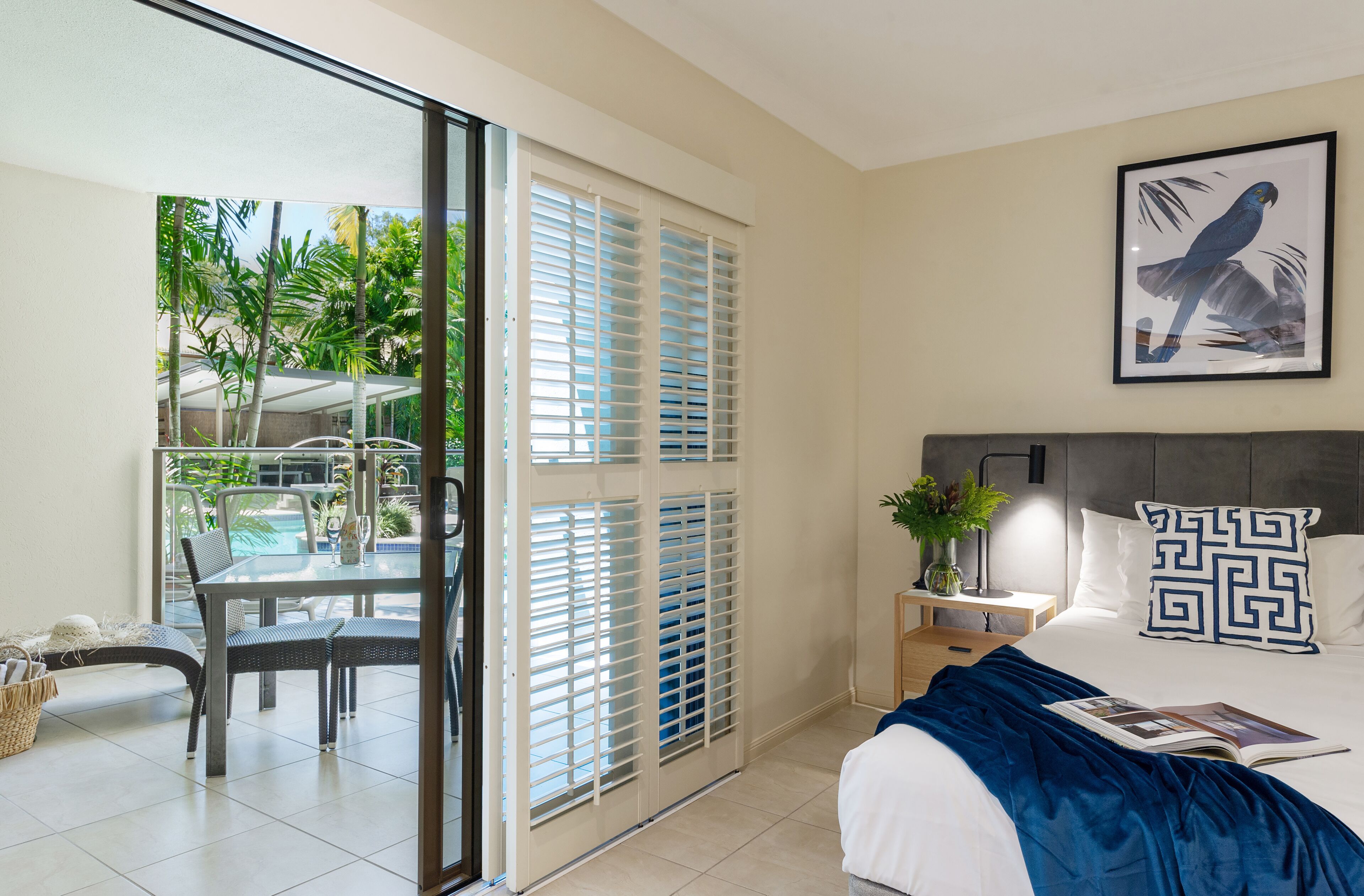 A hotel room with a bed, a nightstand, a framed picture, a window with blinds, and a view of a pool and palm trees.