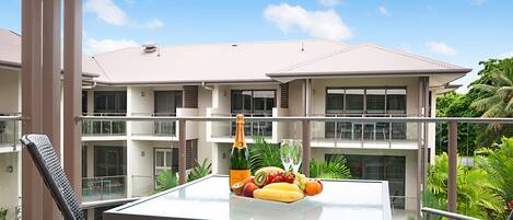 A balcony with a table set for a meal, including fruits and a bottle of champagne, overlooking a residential building and lush greenery.