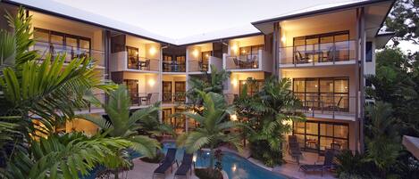 A modern hotel with multiple balconies, a swimming pool, and lush greenery.
