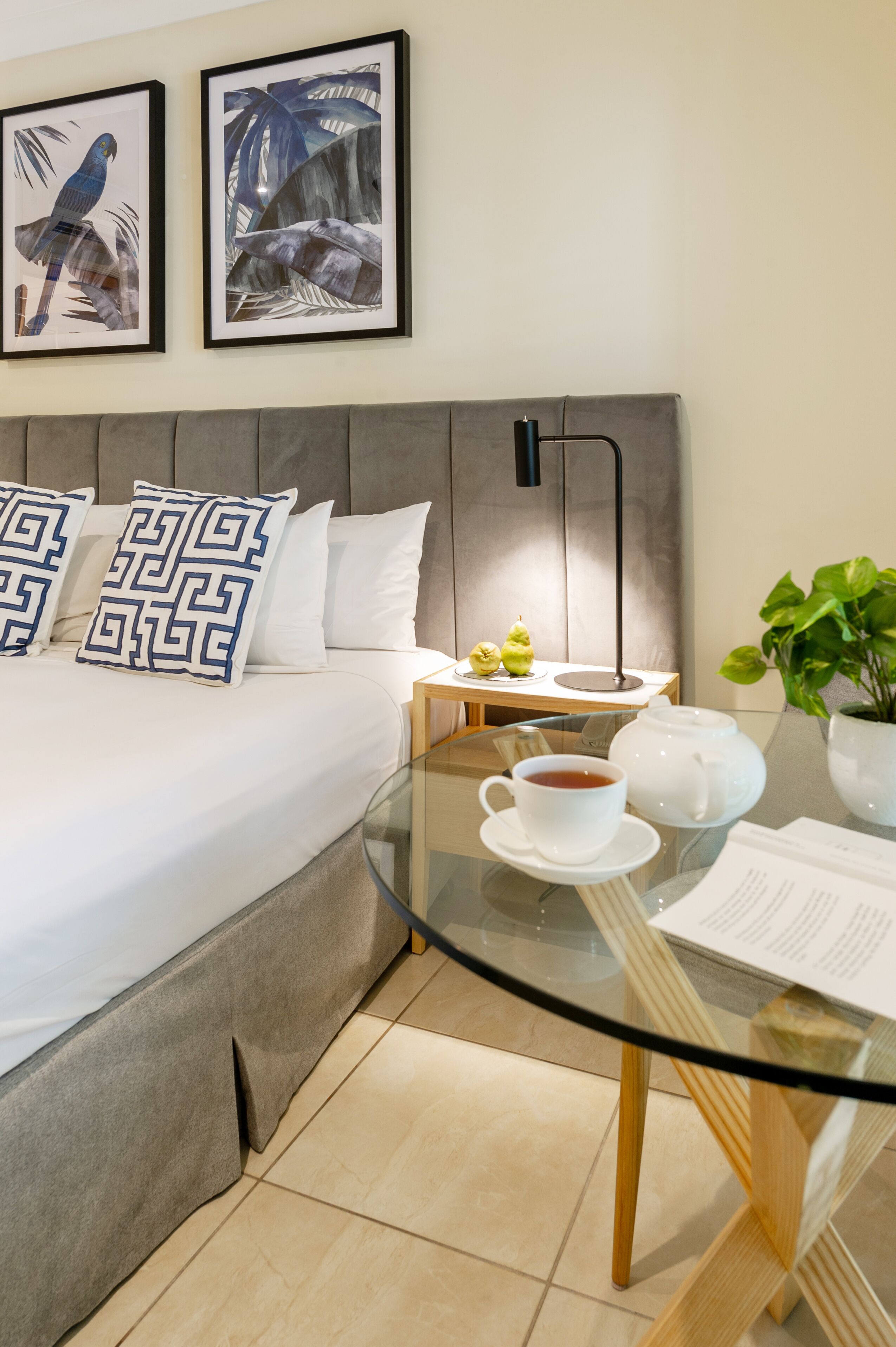 Habitación de hotel ordenada con cama, una mesita de noche con lámpara, una mesa de vidrio con tetera y taza, y dos cuadros en la pared.