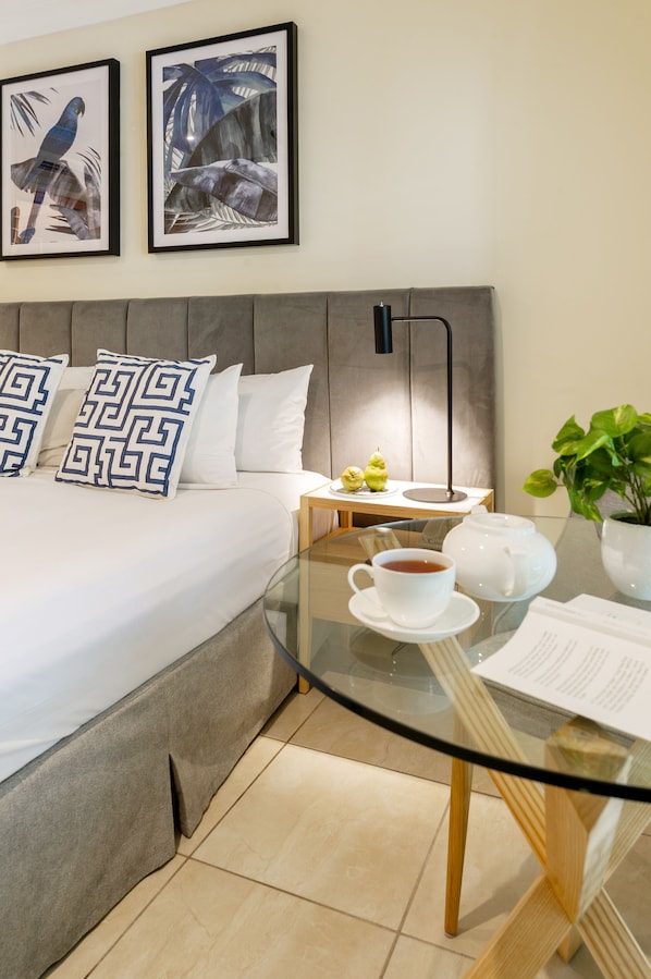 Une chambre d’hôtel bien rangée, avec un lit, une table de chevet dotée d’une lampe, une table en verre avec une théière et une tasse, et deux tableaux encadrés accrochés au mur.