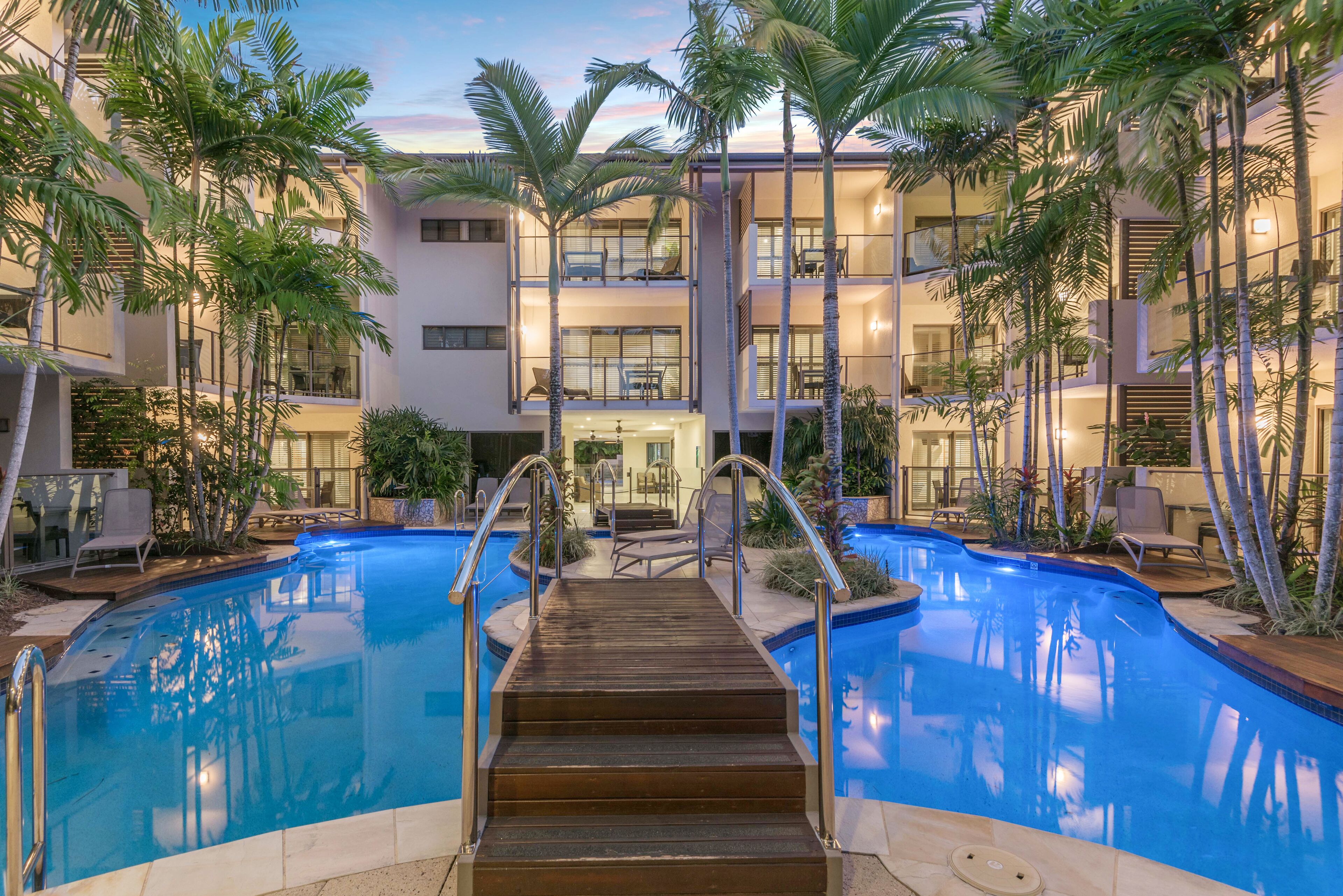 A pool area with a wooden deck leading to the pool, surrounded by palm trees and a building with balconies.
