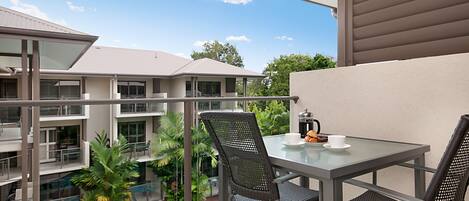 A balcony with a glass table and chairs, overlooking a swimming pool and a building with multiple balconies.