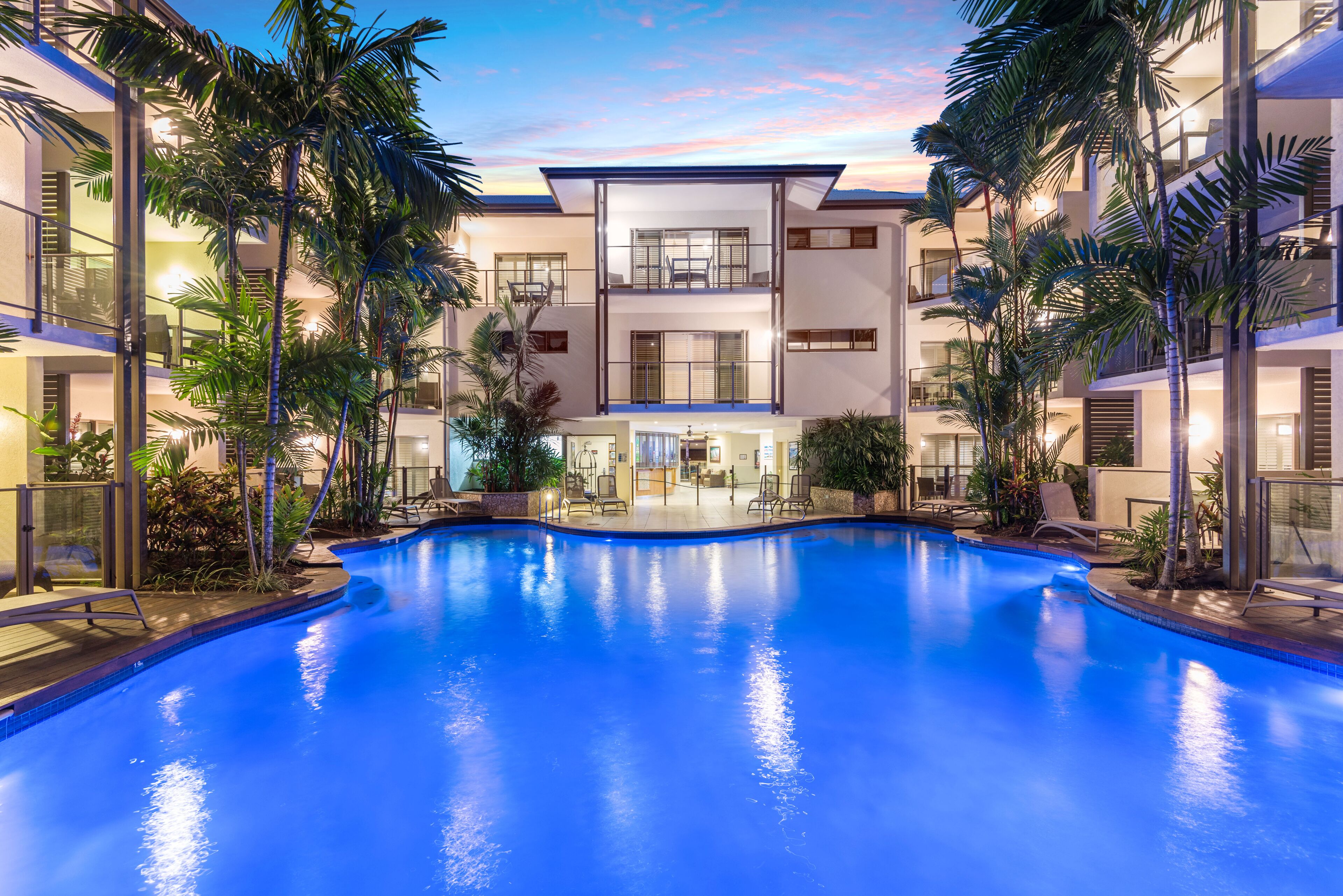 A modern hotel with a central pool, surrounded by palm trees and a multi-story building with balconies.