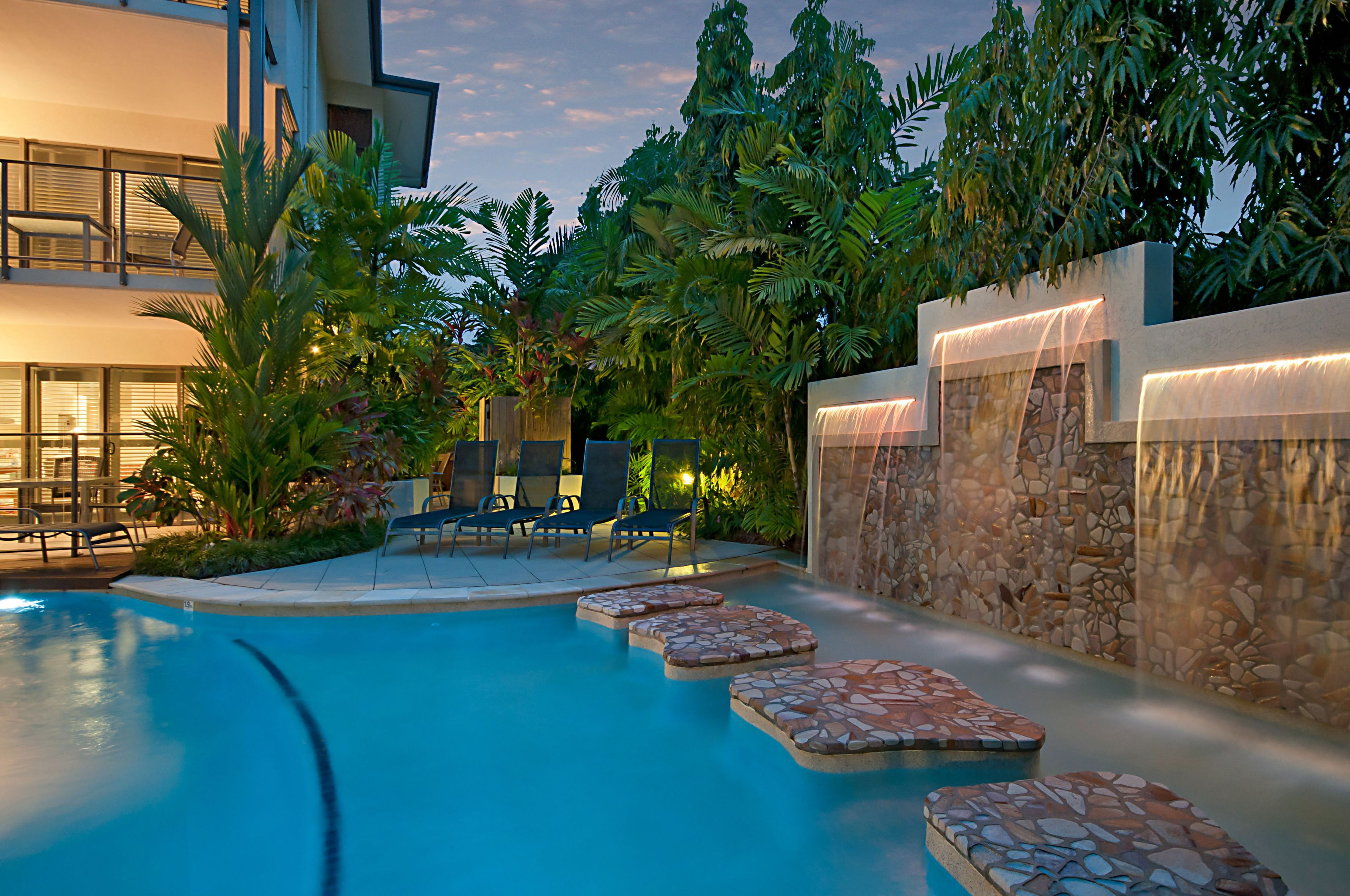 Piscina con sillas de descanso y pared de piedra con fuente de agua.
