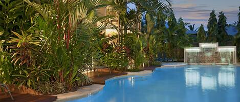 A swimming pool with a wooden deck, surrounded by tropical plants and a stone wall.