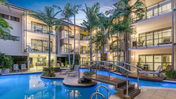 Un hotel de varios pisos con piscina central, mobiliario exterior y palmeras.