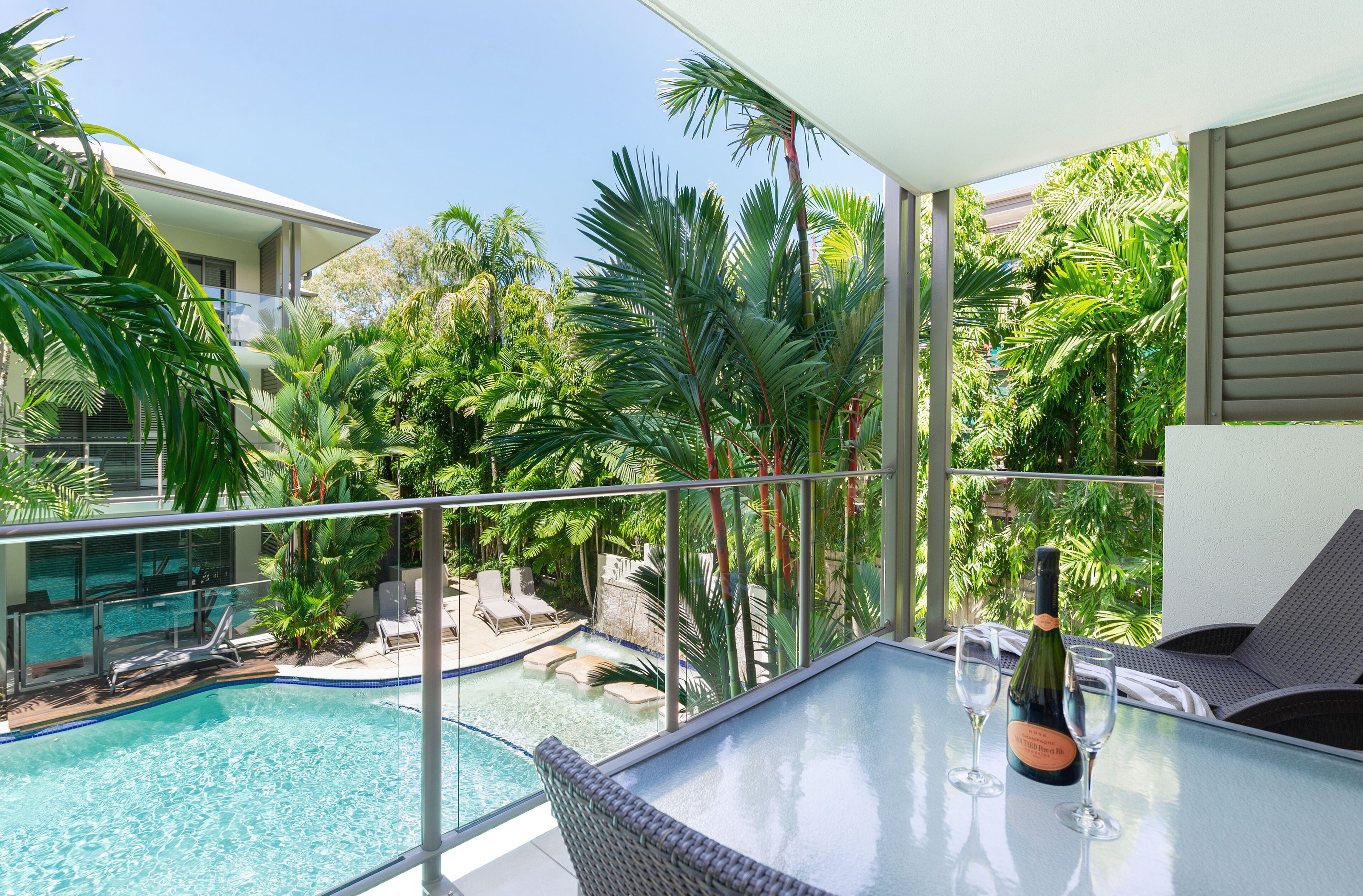 Ein Balkon mit Glastisch, zwei Stühlen, einer Champagnerflasche und einem Swimmingpool mit Liegestühlen.