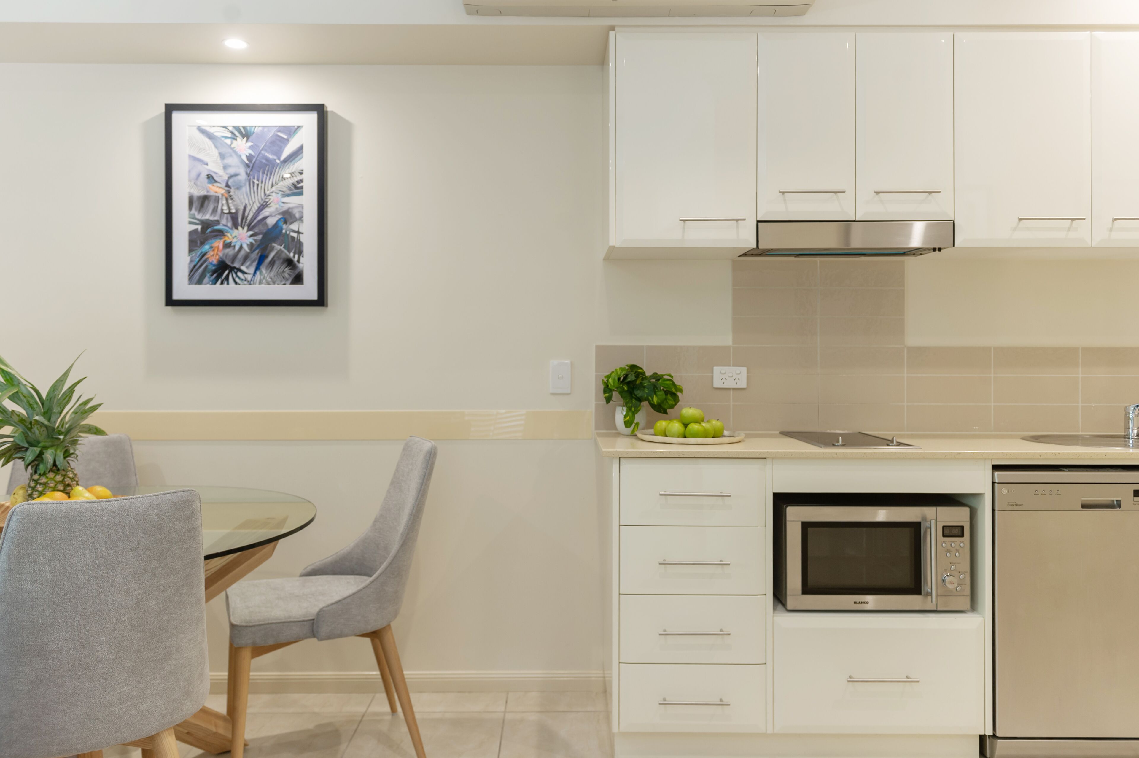 A modern kitchen with white cabinets, a stainless steel microwave, and a dishwasher. There is a dining table with chairs and a framed picture on the wall.