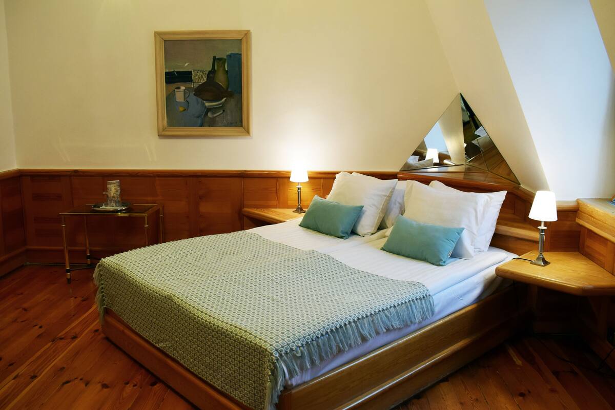 Ein ordentlich bezogenes Bett mit grüner Decke, zwei Nachttische mit Lampen, ein Holz-Kopfteil, ein Bild an der Wand und ein Spiegel, der einen Teil des Raumes reflektiert.