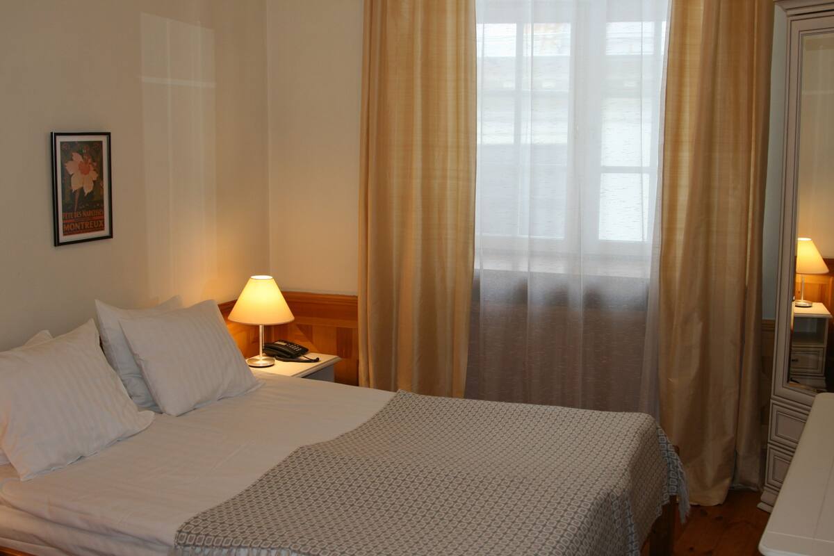Ein Hotelzimmer mit einem ordentlich bezogenen Bett, einer Nachttischlampe, einem gerahmten Bild an der Wand und einem Fenster mit Vorhängen.