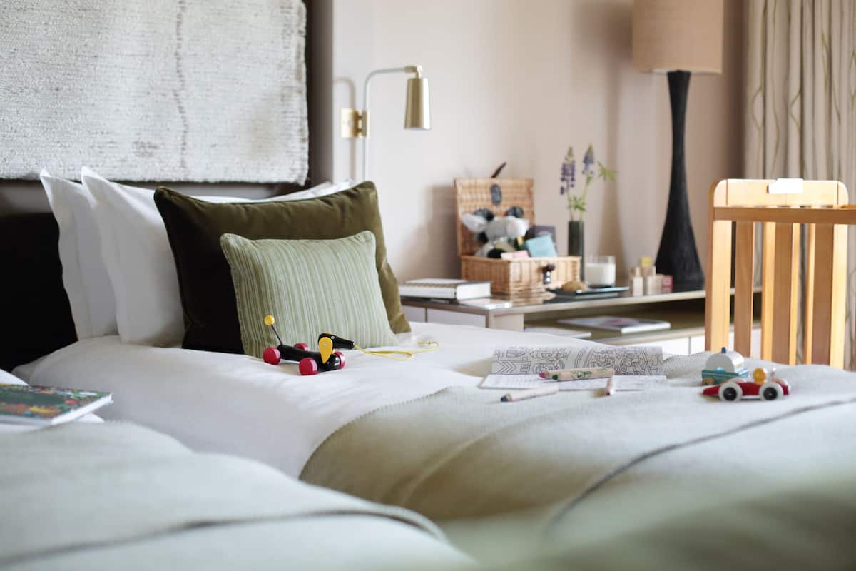 A neatly made bed with pillows, a bedside table with a lamp, a small wooden chair, and a basket of items.