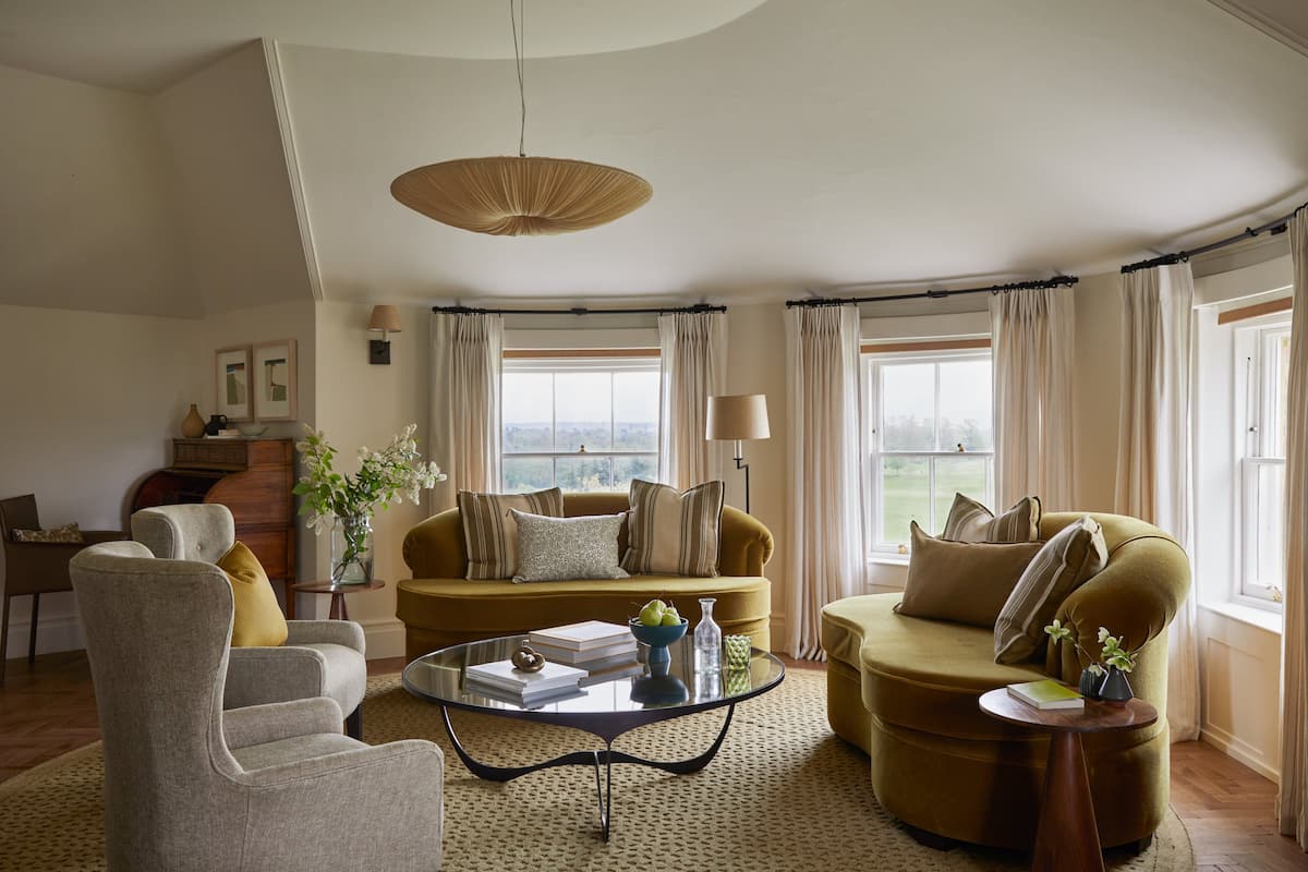 A living room with a sofa, armchair, and coffee table, a lamp, and a view through large windows.