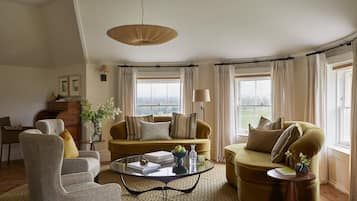 A living room with a sofa, armchair, and coffee table, a lamp, and a view through large windows.