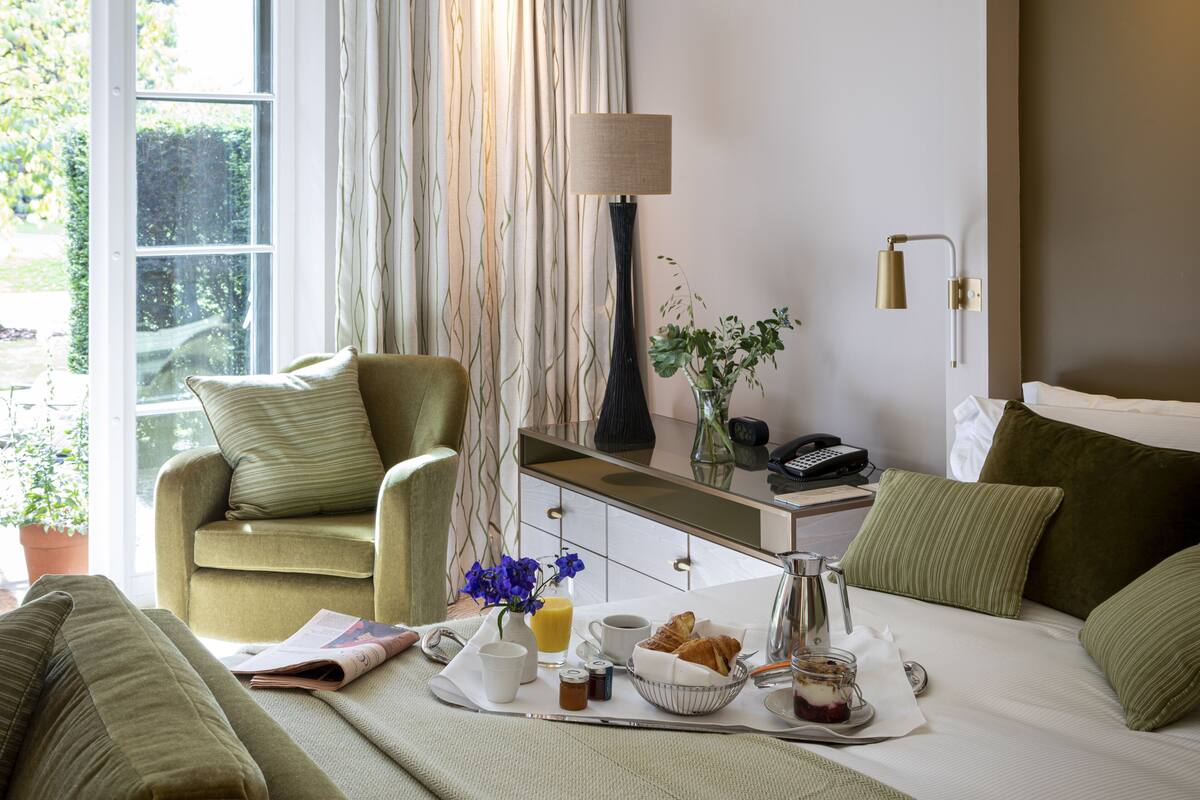 A hotel room with a bed, a sitting area with a chair and a table, a bedside lamp, a vase with flowers, and a breakfast tray.