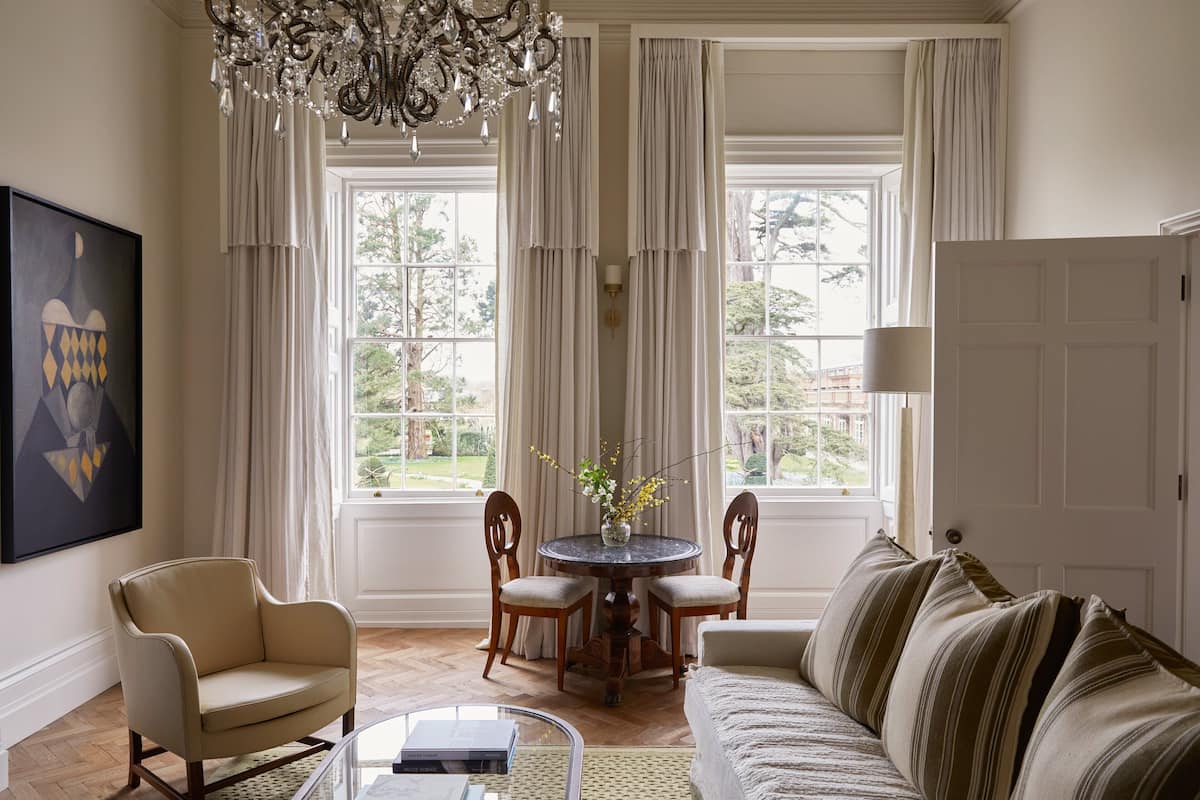 A living room with a beige sofa, a round table with chairs, a chandelier, and large windows with curtains.