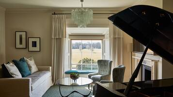 A living room with a sofa, armchairs, a coffee table, a piano, and a view of the outdoors through a large window.