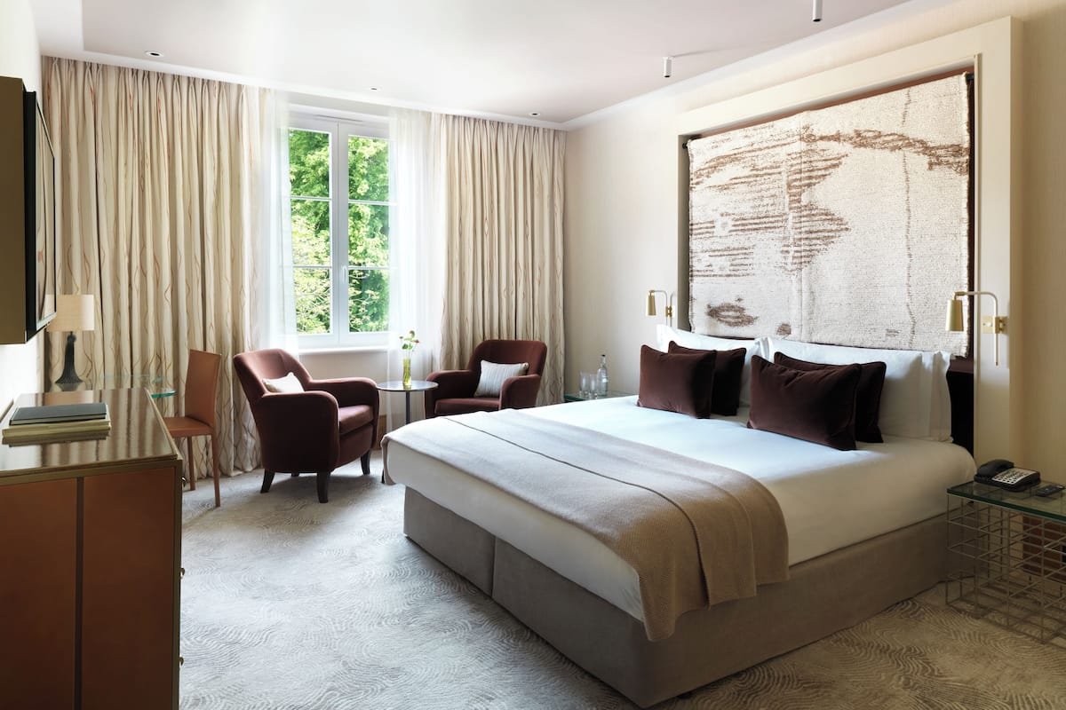A hotel room with a large bed, two armchairs, a small table, and a view of greenery through the window.