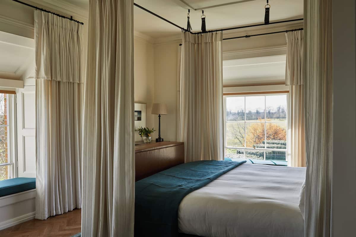 A bedroom with a large bed, a bedside table with a lamp, and a window with curtains.