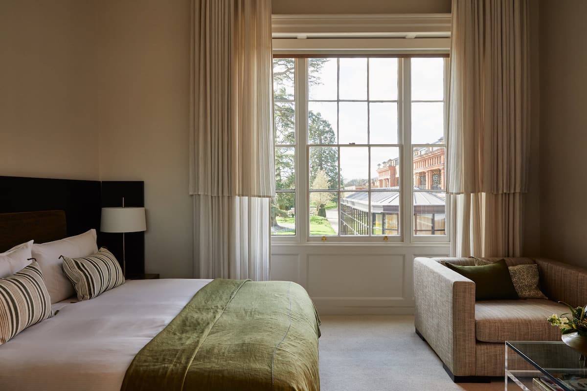 A hotel room with a large bed, a sofa, a glass coffee table, and a view of a building through a window.
