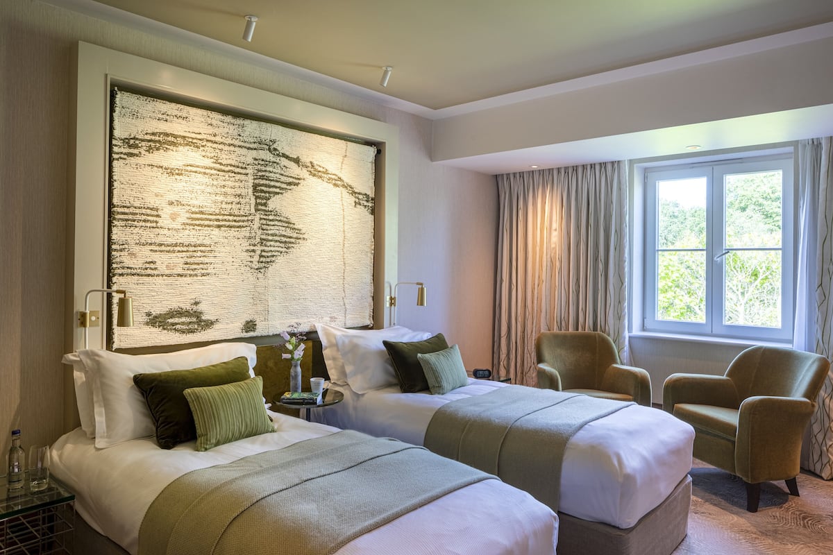 A hotel room with two beds, a large abstract painting on the wall, a window with curtains, and a chair.
