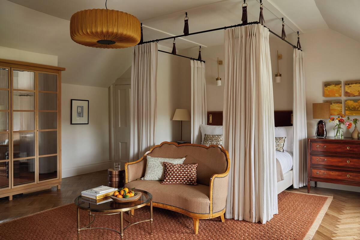 A bedroom with a four-poster canopy bed, a wooden cabinet, a round coffee table with books and fruit, and a large window with curtains.