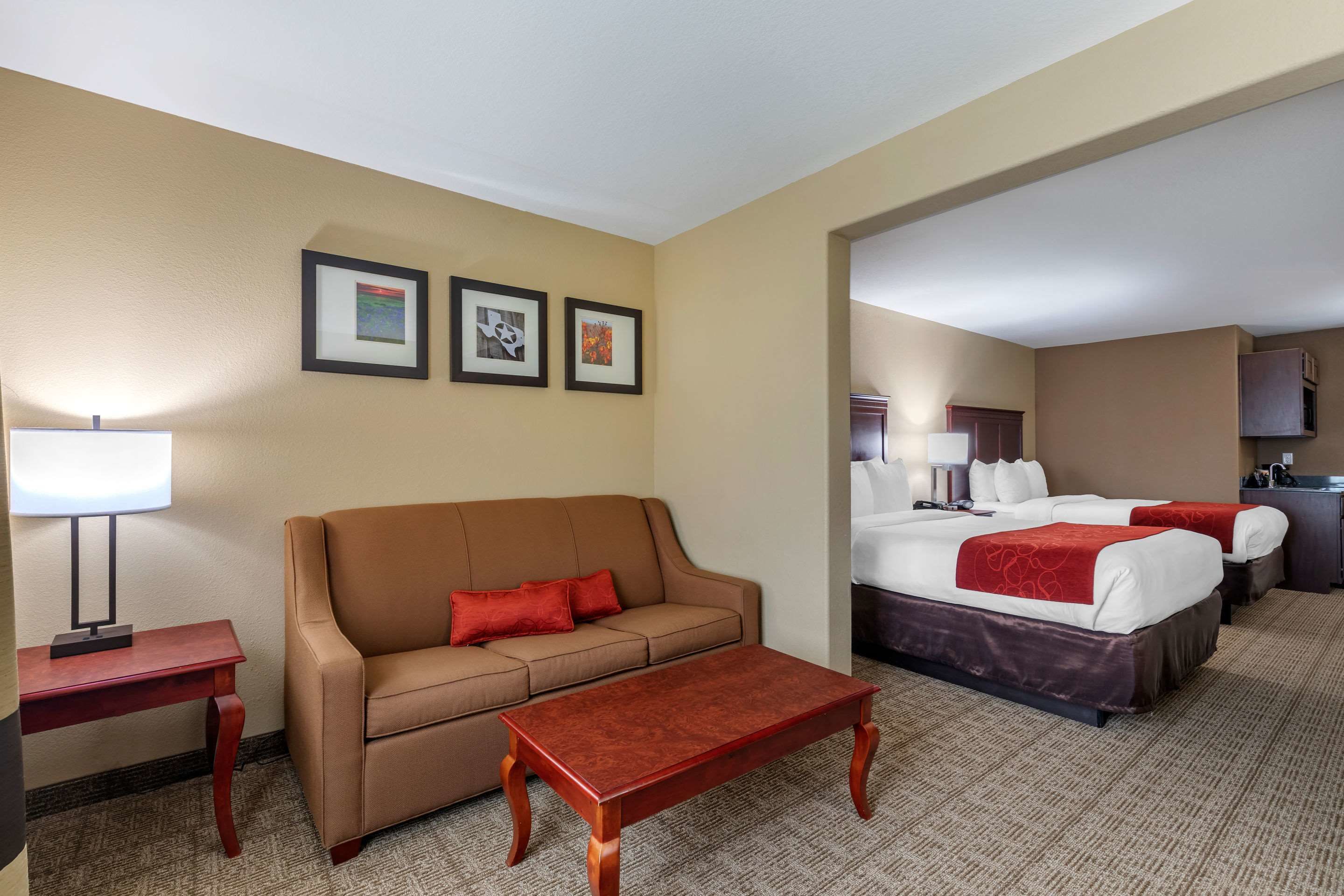 Habitación de hotel con un sofá marrón, una mesa de centro de madera, una cama con ropa de cama blanca y roja, y un escritorio pequeño con una lámpara.