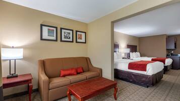 Habitación de hotel con un sofá marrón, una mesa de centro de madera, una cama con ropa de cama blanca y roja, y un escritorio pequeño con una lámpara.