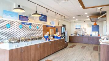 A breakfast area with a counter displaying hot food options, a refrigerator, and a coffee machine.