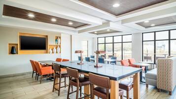 A spacious room with a large table and chairs, a flat-screen TV mounted on the wall, and large windows offering a view of the outdoors.