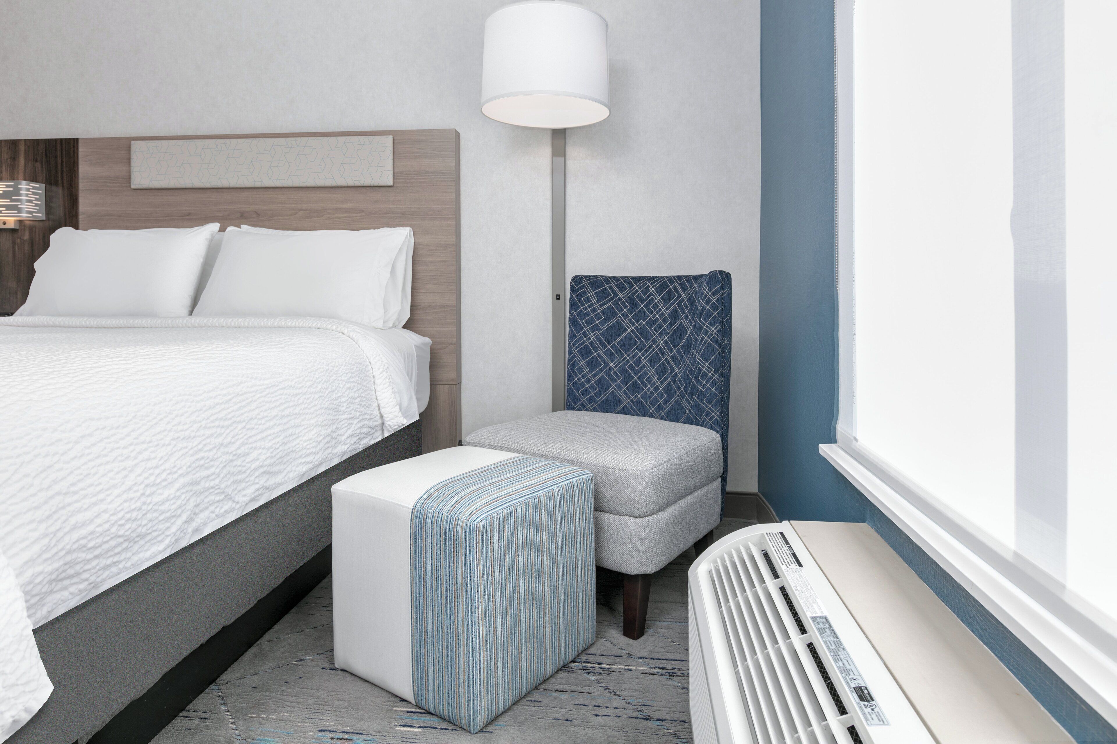 A hotel room with a bed, a bedside table, a lamp, a chair, and a heater.