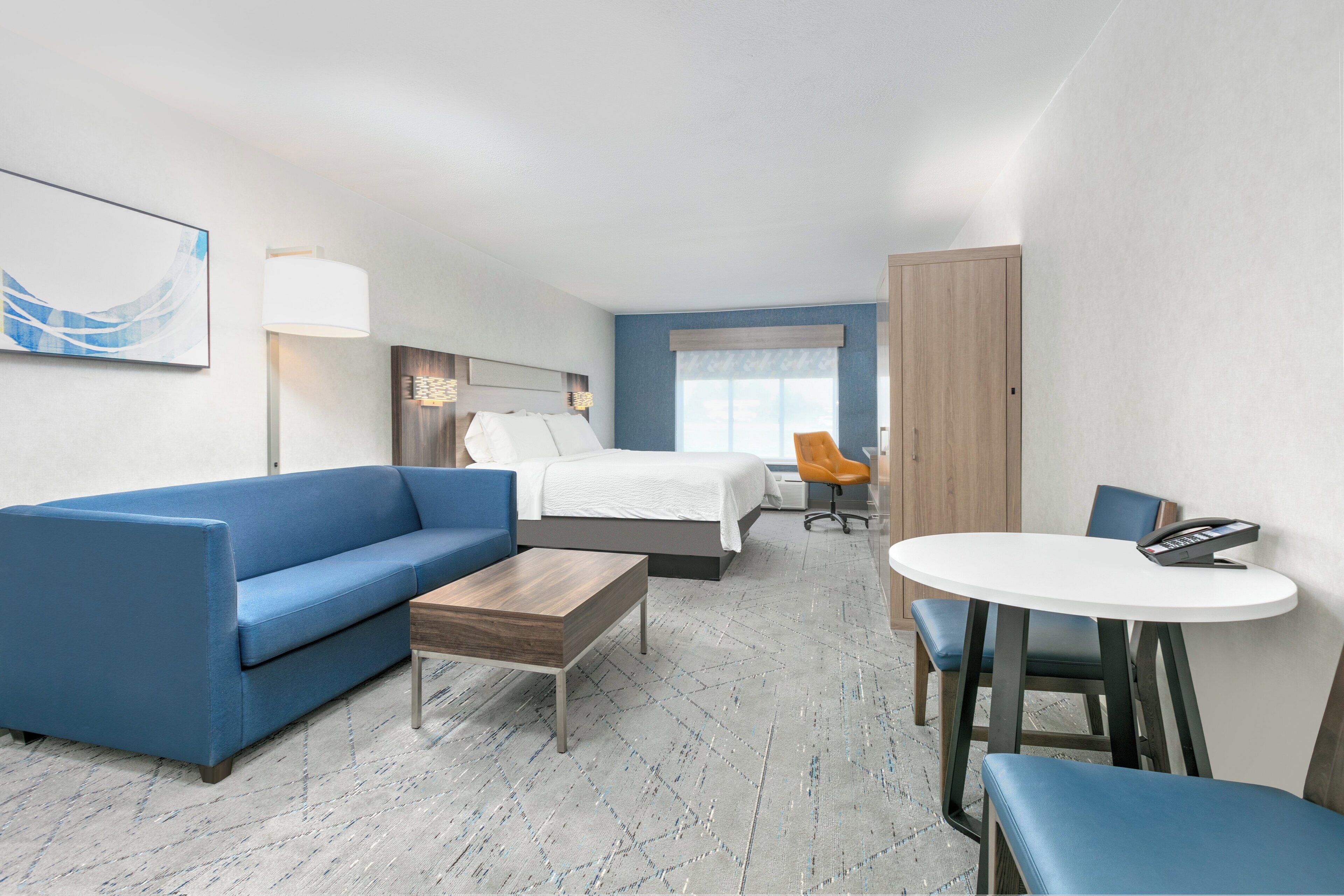 A modern hotel room with a blue sofa, a bed, a small table, and a round dining table with chairs.