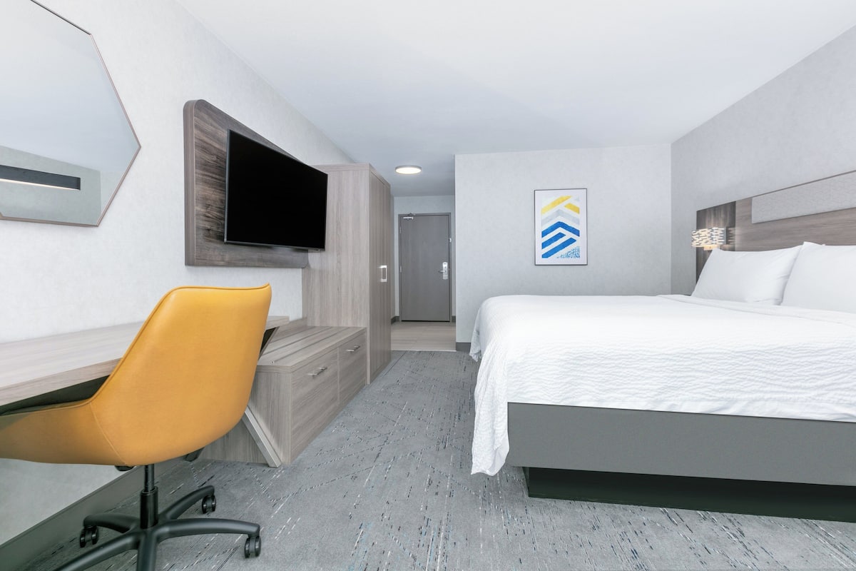 A hotel room with a bed, a desk with a chair, a television mounted on the wall, and a door leading to another room.