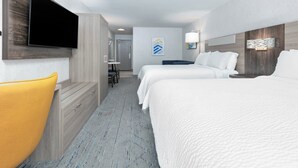 Ein Hotelzimmer mit Wand-Fernseher, zwei Betten mit weißer Bettwäsche, einem gelben Sessel, einem kleinen Tisch und einem Duschbereich.