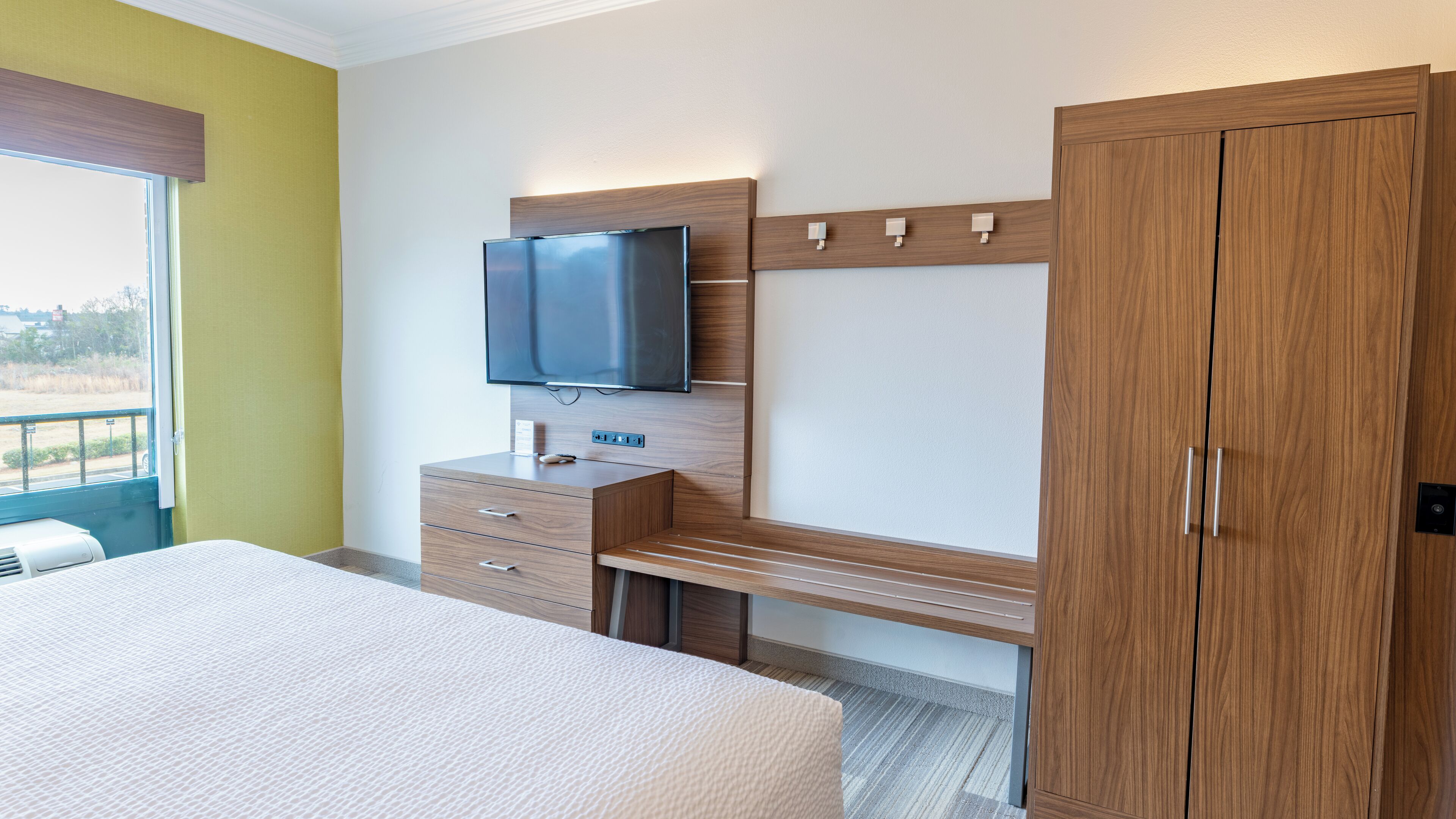 Quarto de hotel com cama, televisão presa a um painel de madeira, guarda-roupa e pequena escrivaninha com gavetas.