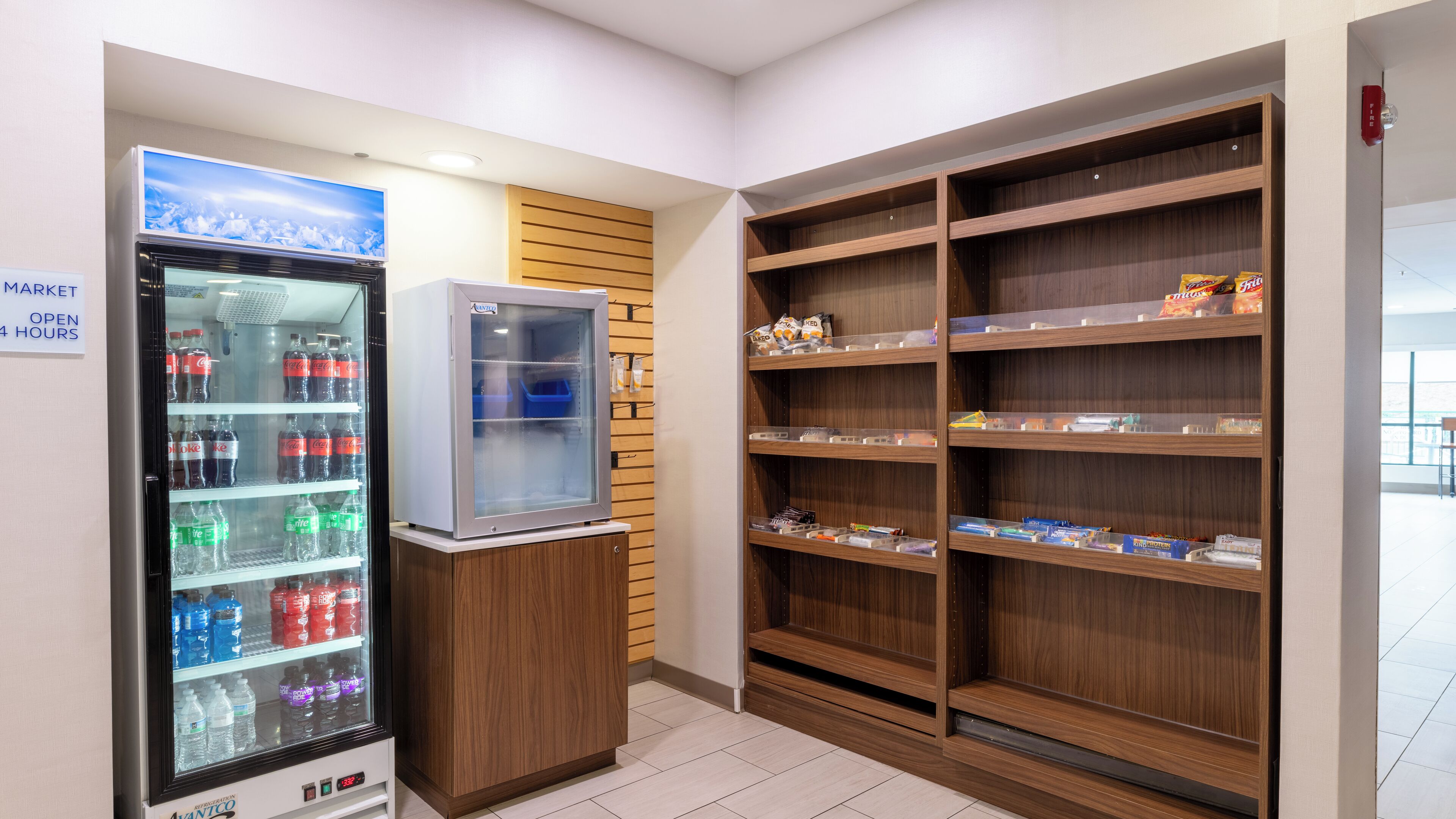 Una máquina expendedora con bebidas y un refrigerador pequeño, estantes de madera con aperitivos, y un extintor en la pared.